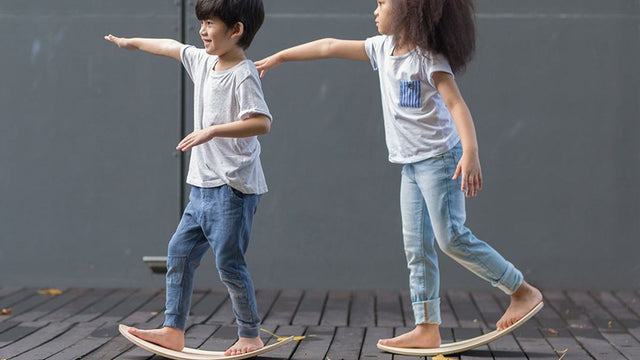 Two children barefoot on curved wooden balance boards with arms outstretched for balance