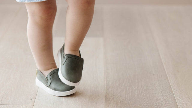 Toddler in a denim skirt mid-step on a pale wood floor wearing sage green slip-on sneakers with white soles