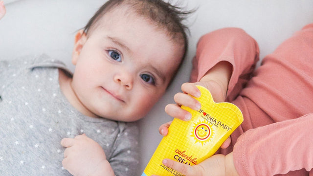 Baby in a grey star-print bodysuit lying on a sofa, holding a yellow tube of California Baby calendula cream