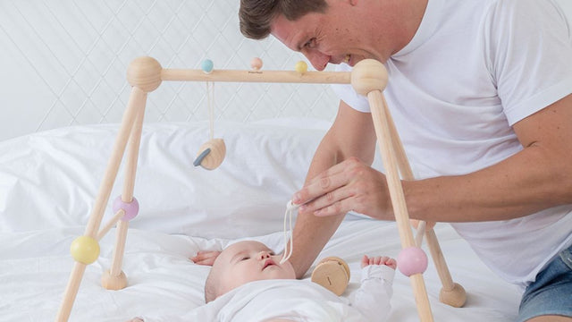 Dad playing with baby under a PLAN Toys wooden play gym made from sustainable rubberwood