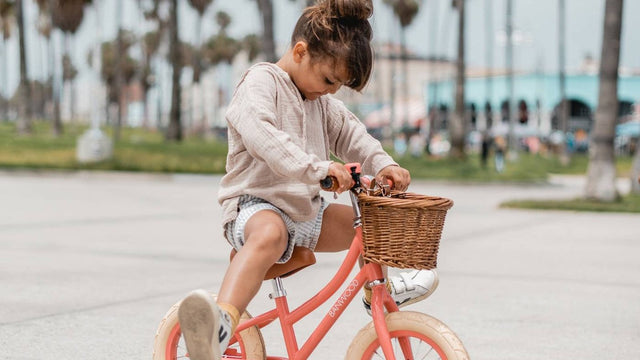 Toddler riding a coral Banwood balance bike with a wicker front basket along a palm-lined seafront path