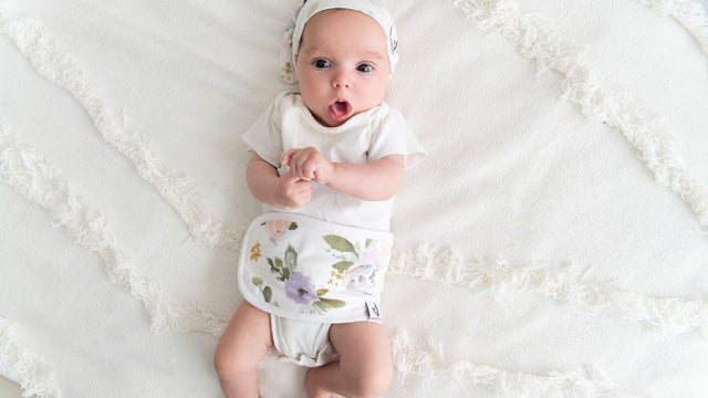 One-month-old newborn in a white outfit lying on a textured bedspread with arms raised
