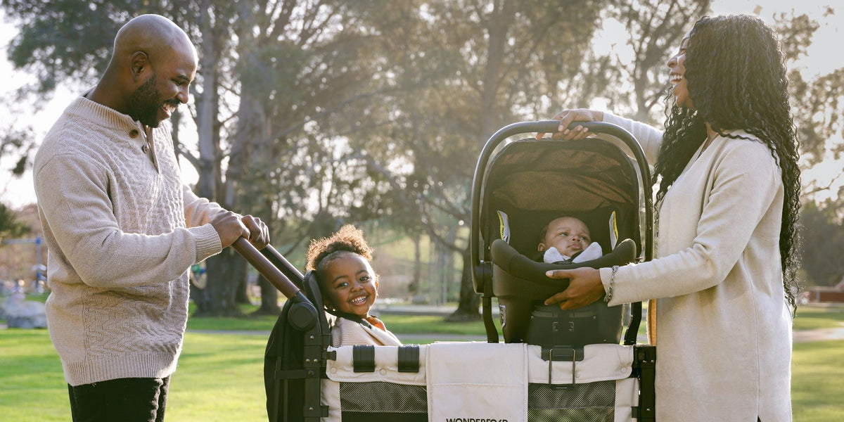 Parents at the park clicking an infant car seat onto a stroller wagon, toddler smiling