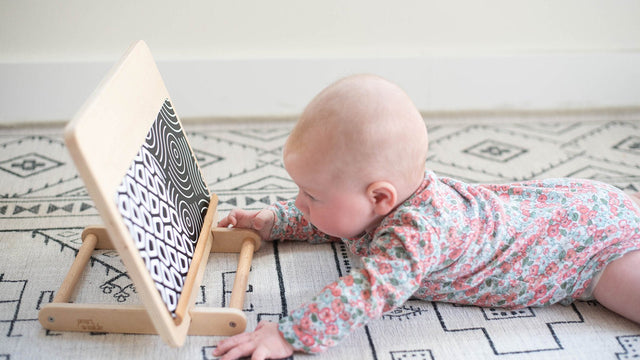 Young baby doing tummy time on a patterned rug, reaching toward a black-and-white high-contrast board