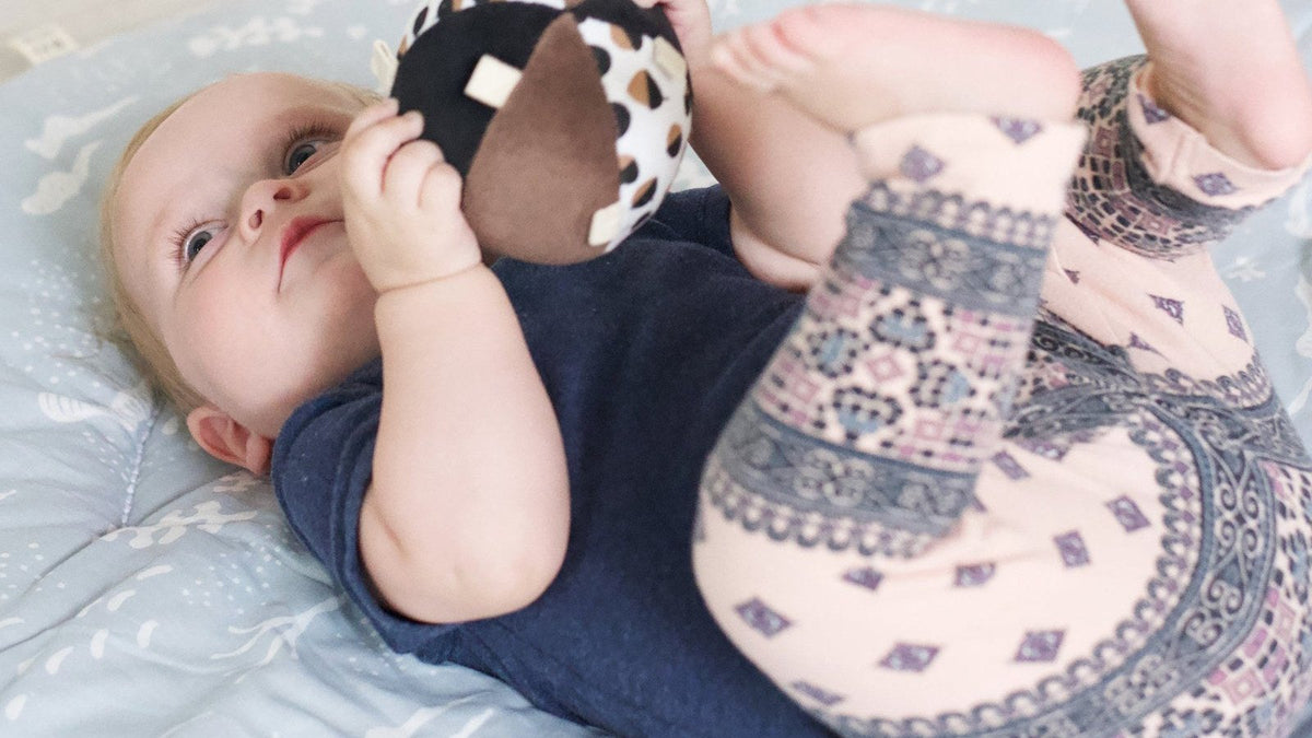 Baby lying on a quilted mat holding a soft black-and-white rattle toy and kicking up both feet