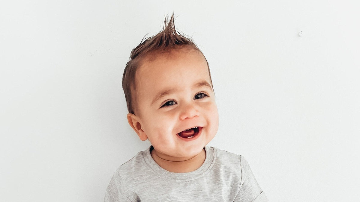Smiling baby with spiky hair showing two new bottom teeth, sitting against a white wall