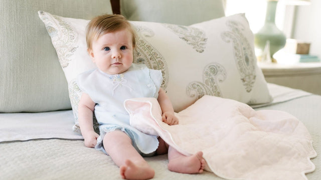 Baby sitting up on a bed propped against a paisley pillow with a quilted blanket over her lap