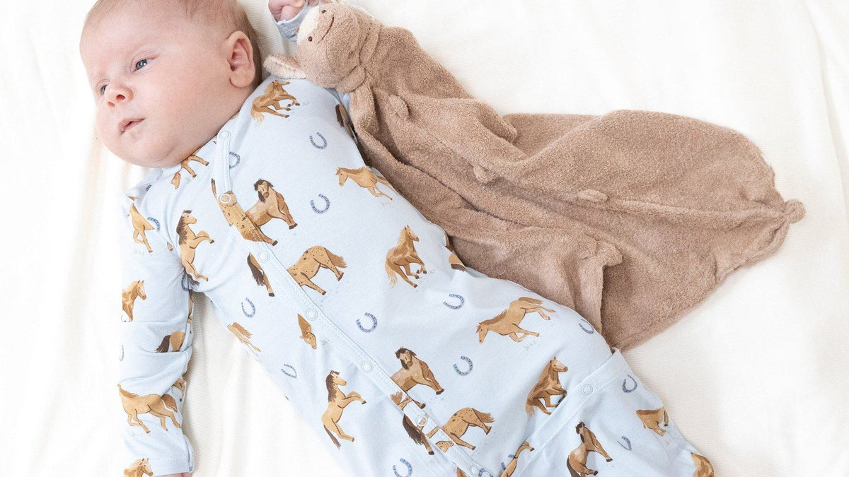 Newborn baby lying on a white bed in an Angel Dear bamboo horse-print gown next to a tan lovey blanket