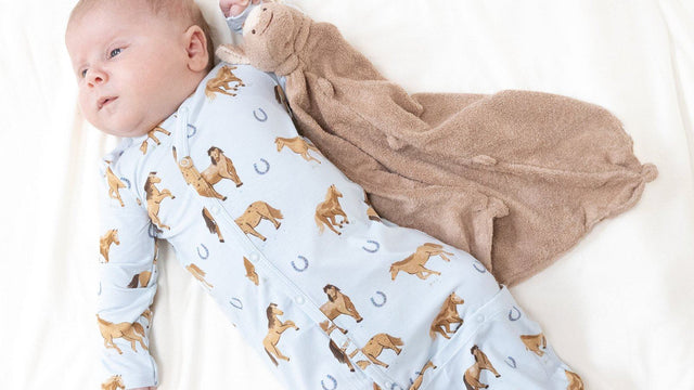 Newborn baby lying on a white bed in an Angel Dear bamboo horse-print gown next to a tan lovey blanket