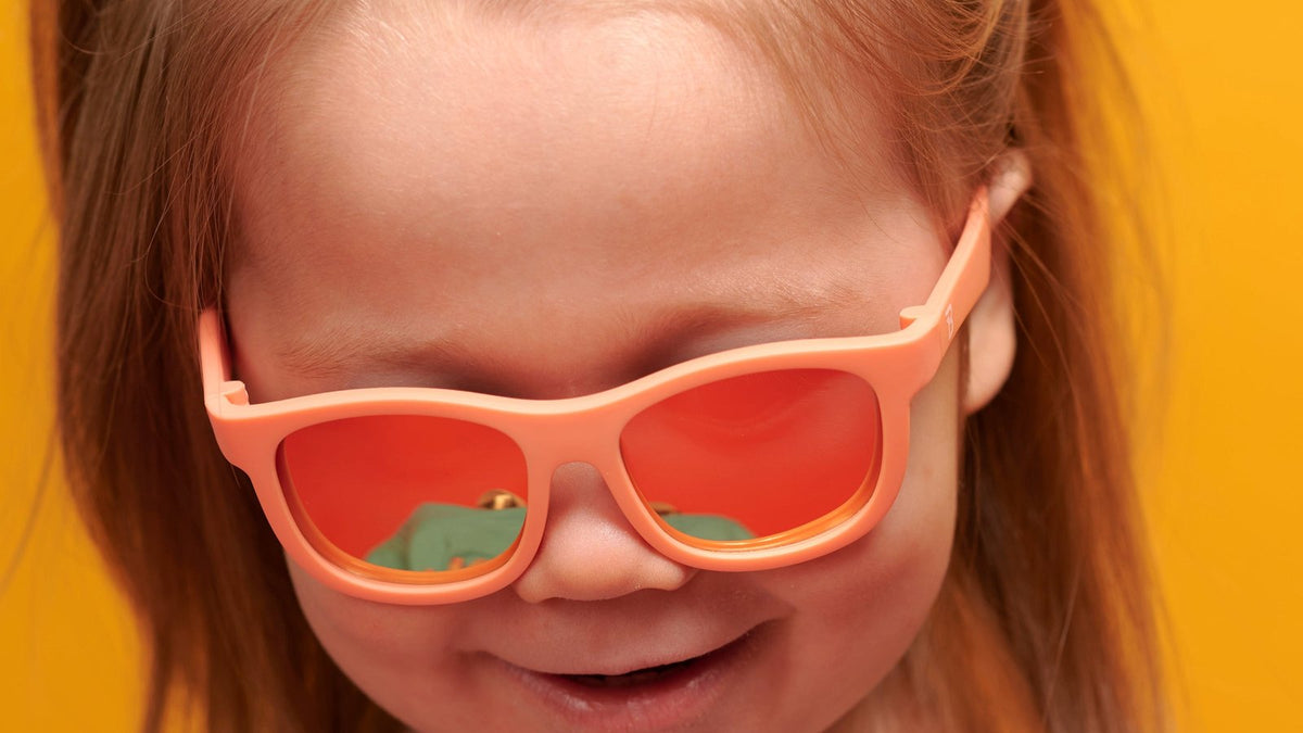 Little girl grinning in papaya-orange Babiators Navigator sunglasses against a yellow backdrop