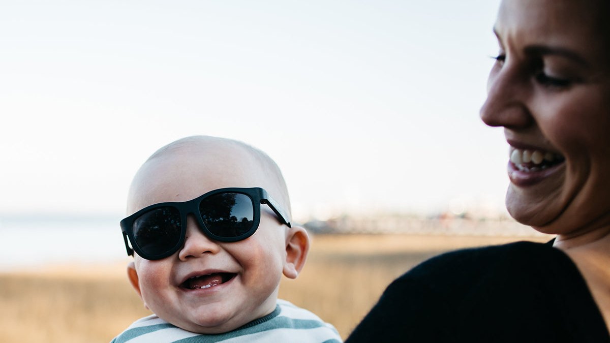 Laughing baby wearing black Babiators Navigator sunglasses held by smiling mother outdoors