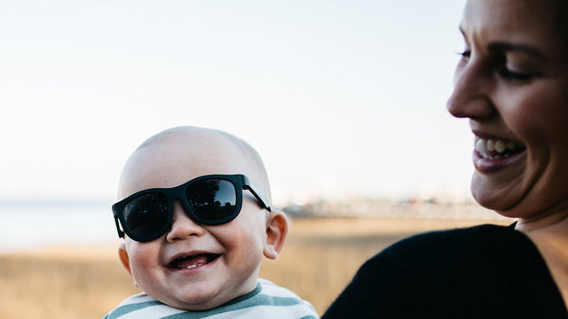 Laughing baby wearing black Babiators Navigator sunglasses held by smiling mother outdoors