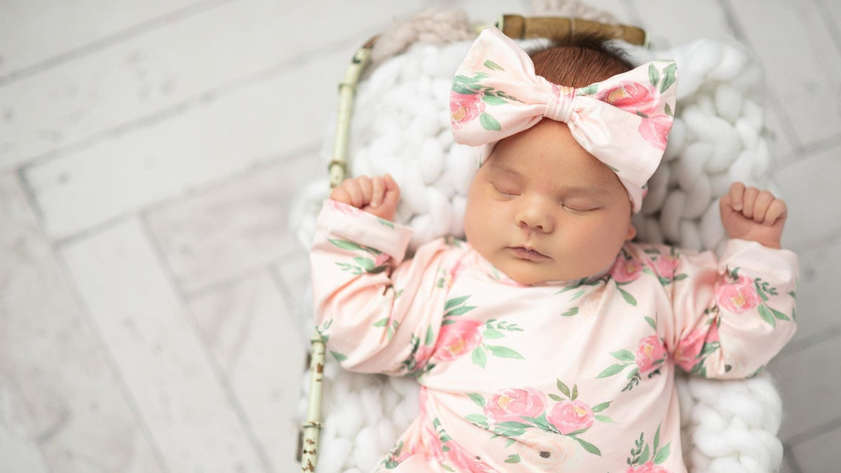 Newborn asleep in a wicker basket on a chunky white knit blanket, wearing pink floral pajamas and a matching bow headband