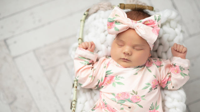 Newborn asleep in a wicker basket on a chunky white knit blanket, wearing pink floral pajamas and a matching bow headband