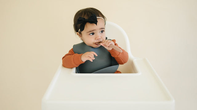 Baby girl in a high chair with a silicone catch-all bib, feeding herself with a soft silicone spoon