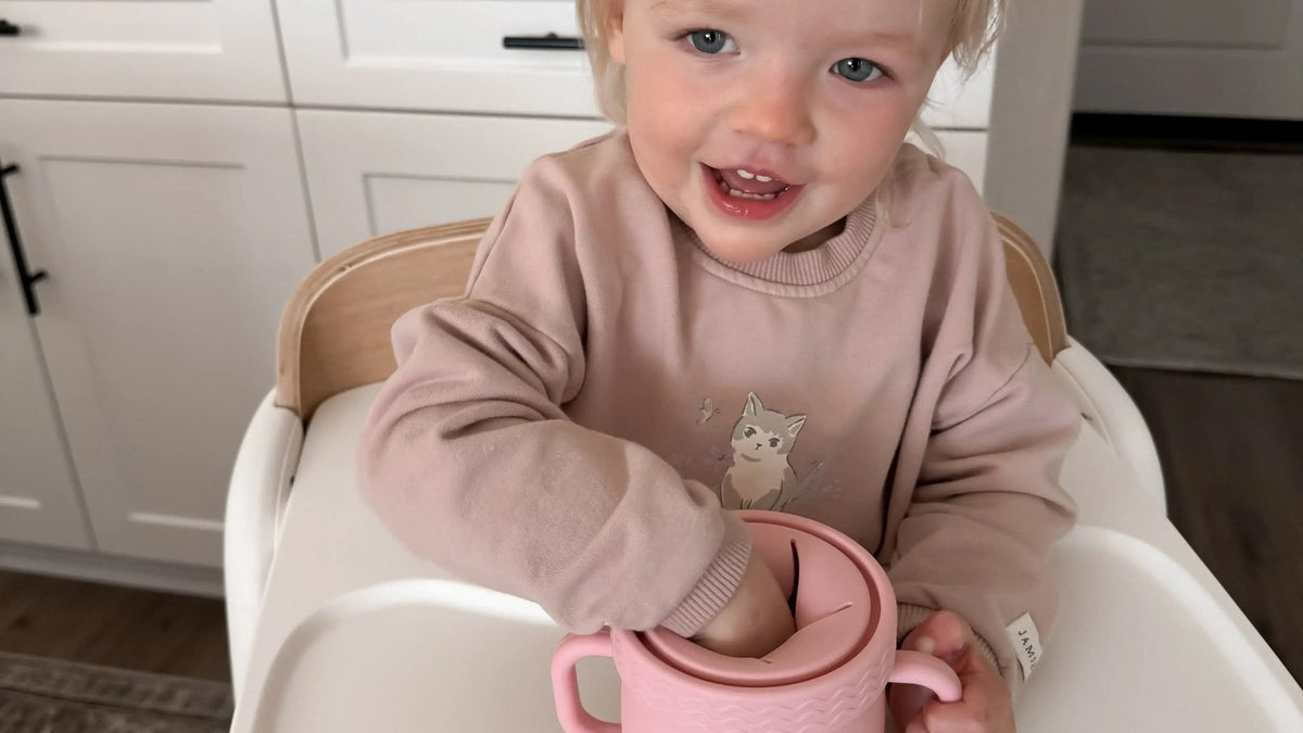 Smiling toddler in a high chair in a white kitchen reaching into a pink silicone snack cup to feed herself