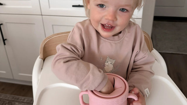 Smiling toddler in a high chair in a white kitchen reaching into a pink silicone snack cup to feed herself