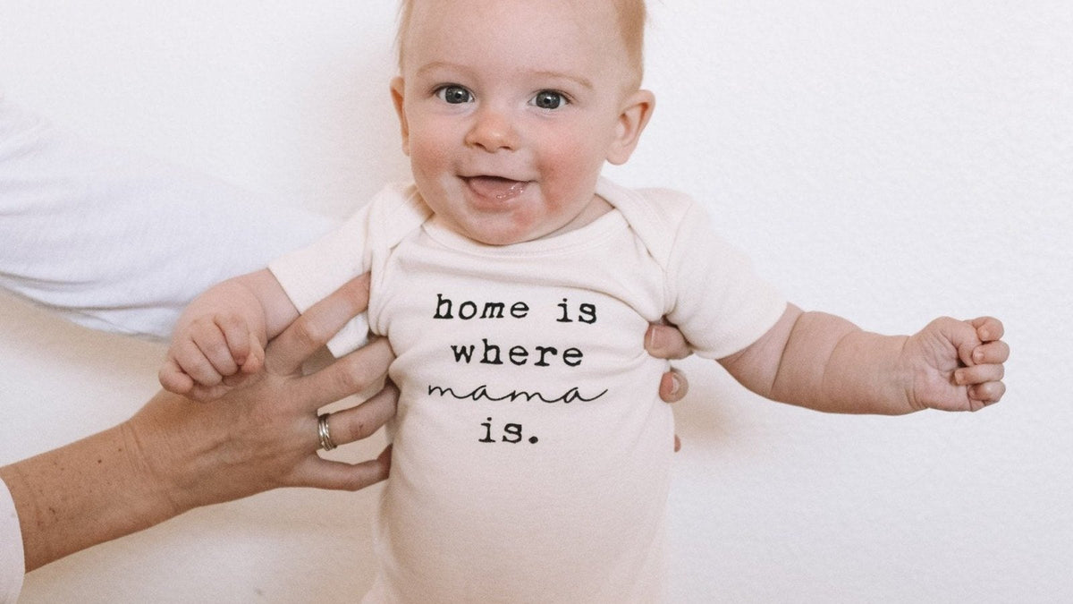 Smiling baby in a short-sleeve onesie printed "home is where mama is", steadied by a parent's hands