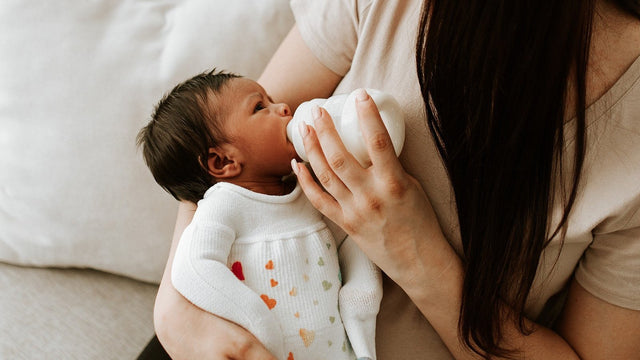 Newborn in a knit outfit being bottle-fed in a parent's arms on the sofa