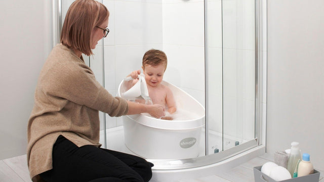 A parent kneeling beside a baby bath tub with bath products in a basket nearby