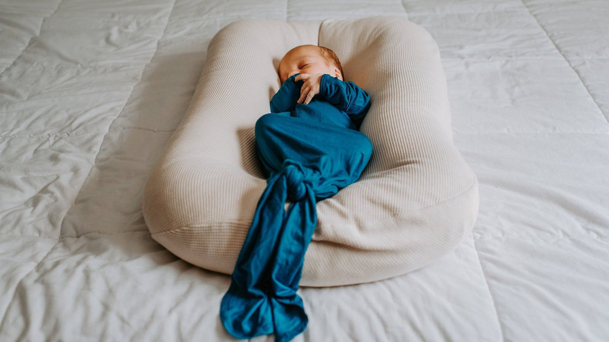 Newborn sleeping peacefully in a teal knotted gown, nestled in a cream baby lounger on a bed