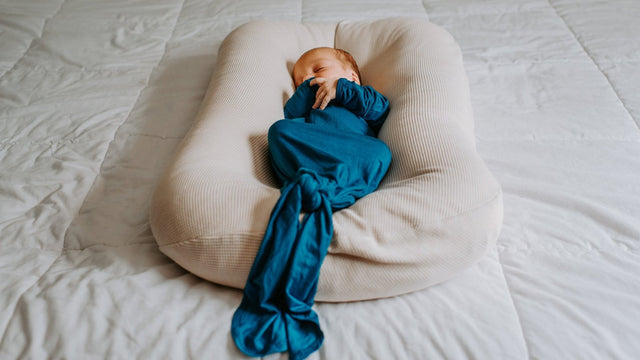 Newborn sleeping peacefully in a teal knotted gown, nestled in a cream baby lounger on a bed