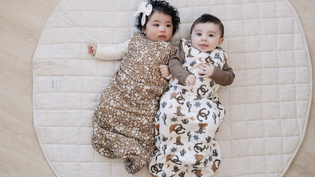 Two babies lying on their backs in floral and western-print wearable sleep sacks on a quilted mat