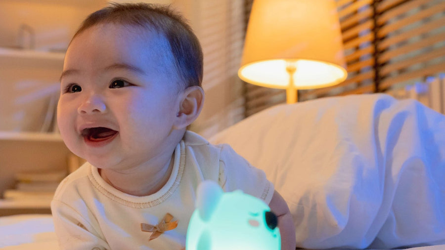 Smiling baby lying on a bed at night beside a softly glowing koala night-light soother, bedside lamp warm in the background