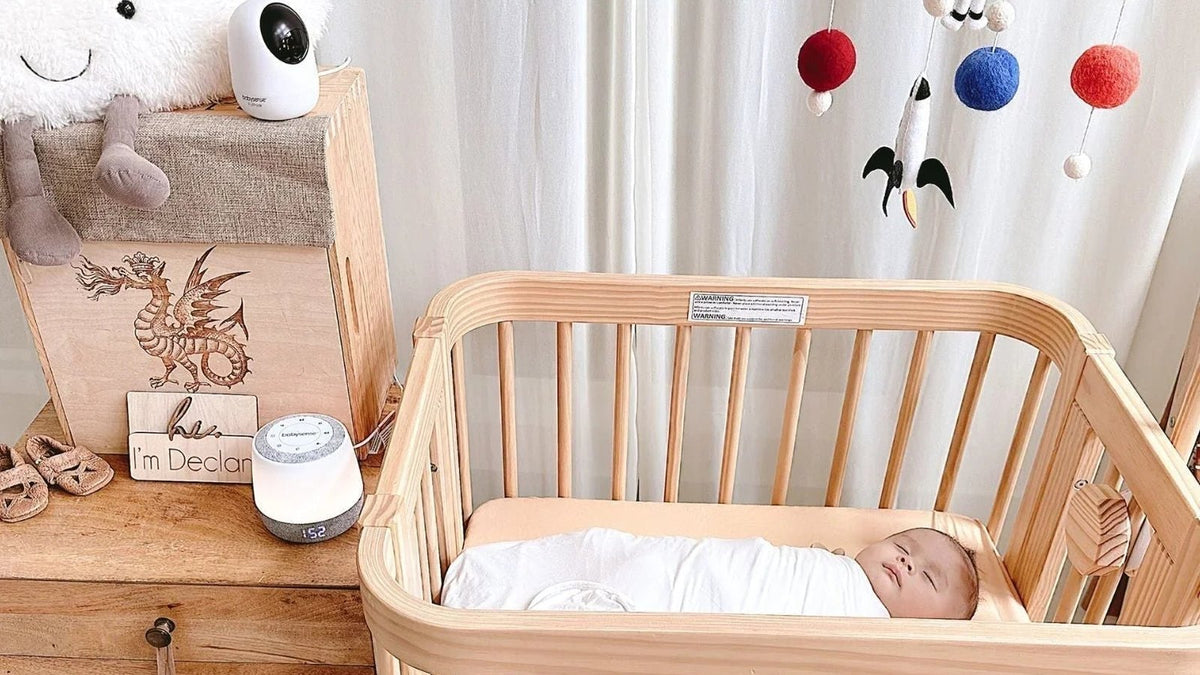 Swaddled newborn asleep in a wooden crib with a Babysense video camera and sound machine on the dresser beside it