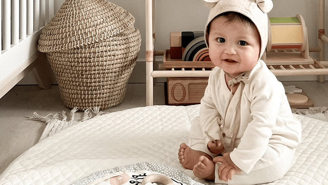 Baby in a bear-ear bonnet sitting on a quilted play mat in a nursery with wooden toys on the shelf behind