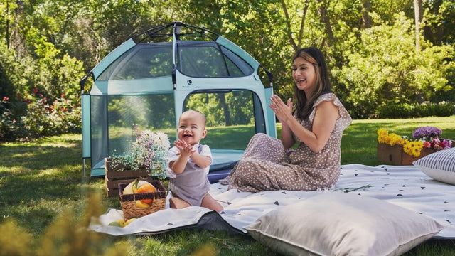 Mom and baby playing on a picnic blanket beside a pop-up beach playpen outdoors
