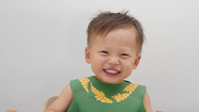 Grinning toddler in a green silicone bib with a yellow lei print, sitting at a high chair tray with a divided plate
