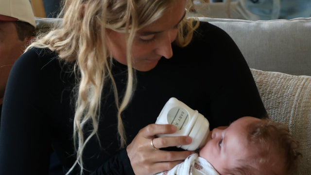 Mother sitting on a sofa bottle-feeding her swaddled newborn from a small Baby K glass bottle