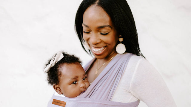 Smiling mother looking down at her baby girl, who is tucked against her chest in a lavender stretchy wrap carrier