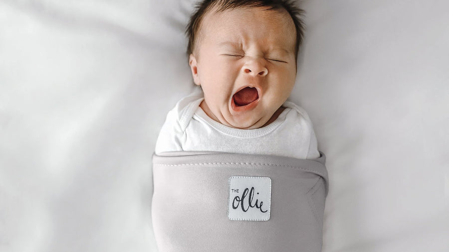 Yawning newborn wrapped snugly in a grey Ollie swaddle on a white sheet