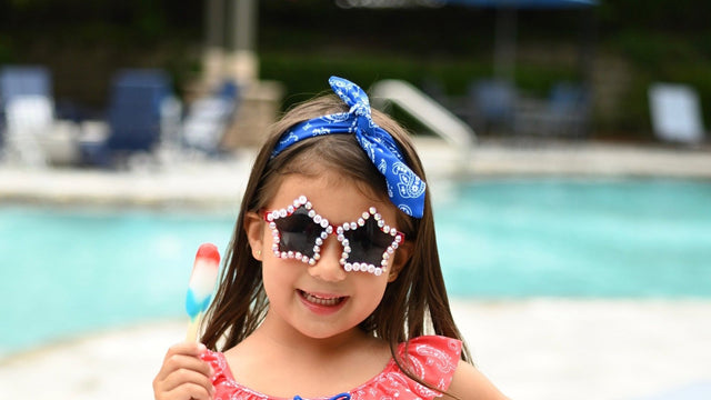 Girl in a red bandana-print ruffle swimsuit, star sunglasses and a blue headband holding a popsicle beside a swimming pool