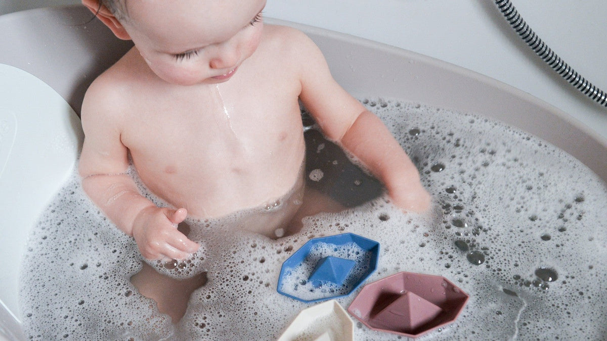 A baby sitting in a bubble bath playing with three floating origami-style bath boats