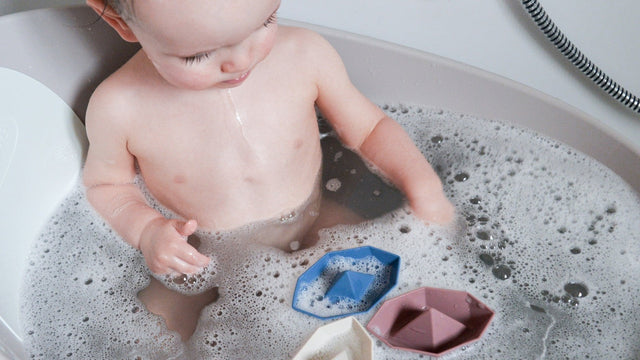 A baby sitting in a bubble bath playing with three floating origami-style bath boats