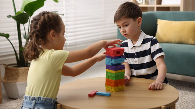 Two children stacking a rainbow wooden block tower on a low round table in a bright living room