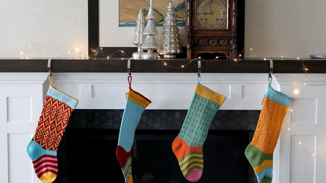 Four colorful knit Christmas stockings hanging from a white mantel strung with fairy lights, with mercury-glass trees and an antique clock above