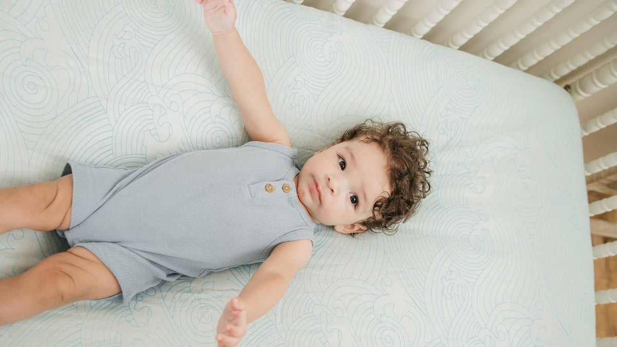 Baby in a blue ribbed romper lying on a white crib mattress fitted with a pale blue sea-waves print bamboo crib sheet, seen from above