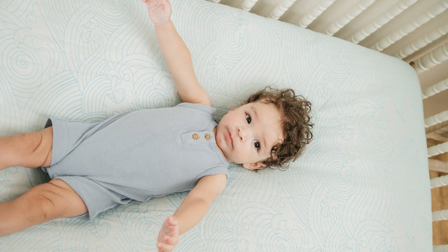 Baby in a blue ribbed romper lying on a white crib mattress fitted with a pale blue sea-waves print bamboo crib sheet, seen from above