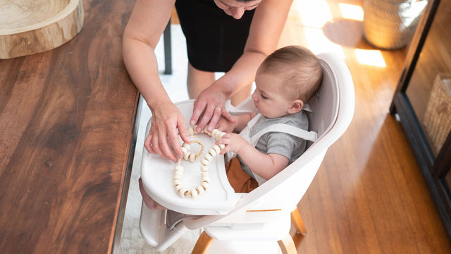Parent leaning over a wooden dining table to hand a baby a teething toy while the baby sits harnessed in a white Trubliss Turn-a-Tot high chair
