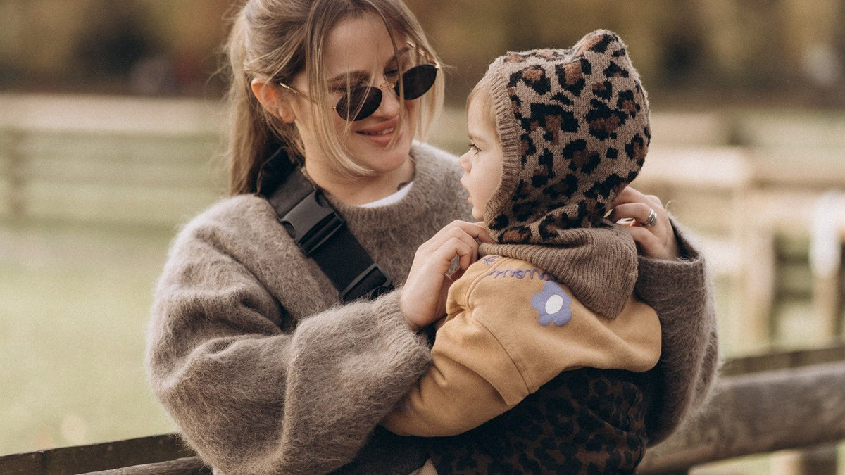 Caregiver carrying a toddler on the hip outdoors with a baby carrier strap over one shoulder