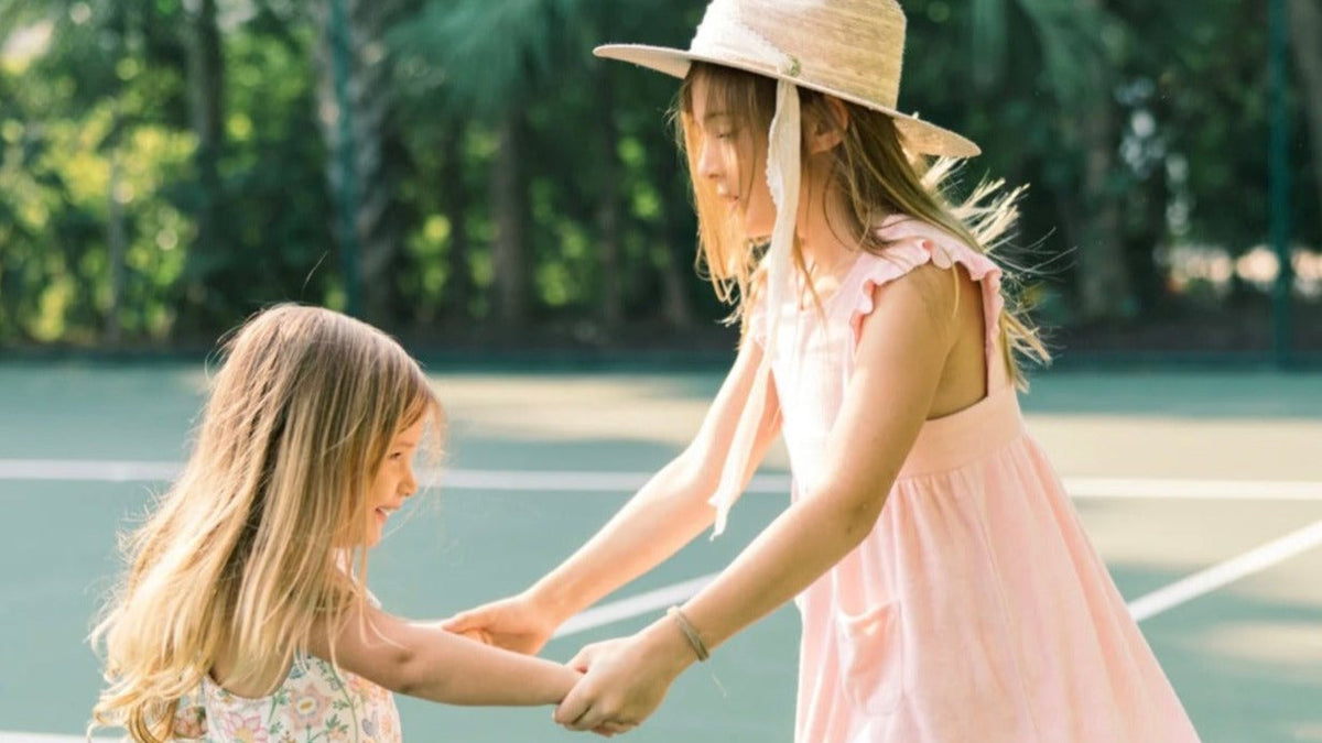 Two girls playing outside, the older one wearing a wide-brim straw kids sun hat