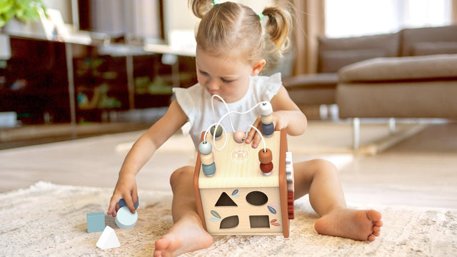 Toddler sitting on a rug fitting wooden shapes into a Montessori 5-in-1 activity cube
