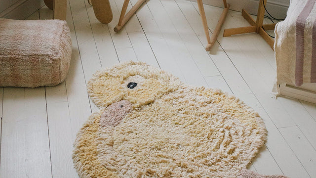 Cream duckling-shaped wool nursery rug on white painted floorboards beside a bassinet stand, wooden stool and blush floor pouf