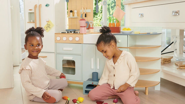 Two girls playing with wooden play food in front of a large wooden play kitchen