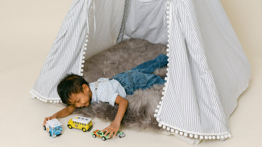 Toddler lying on a sheepskin inside a striped pom-pom play teepee, rolling toy cars through the doorway