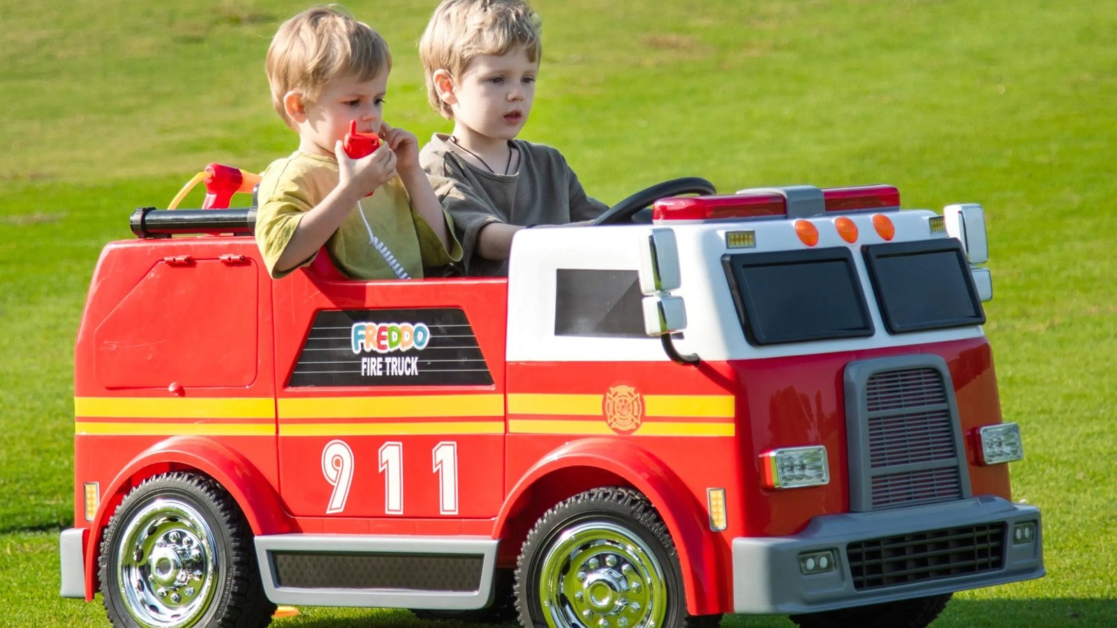 Two children riding a 24V electric ride-on fire truck on a lawn